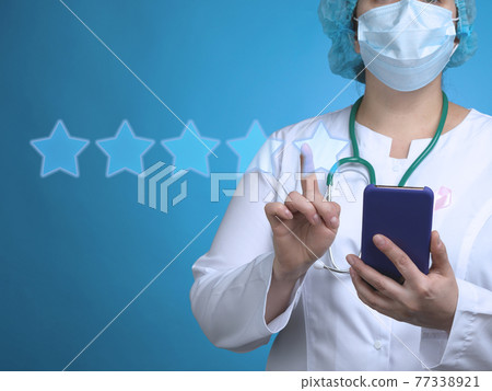 A woman doctor stands on a blue background and holds a smartphone, with the other hand clicks on the virtual rating stars A woman doctor stands on a blue background and holds a smartphone, with the other hand clicks on the virtual rating stars 77338921