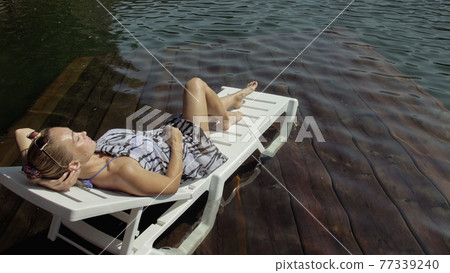 Woman lie on a sunbed in sunglasses and a boho silk shawl. Girl rest on a flood wood underwater pier. The pavement is covered with water in the lake. Woman lie on a sunbed in sunglasses and a boho silk shawl. Girl rest on a flood wood underwater pier. The pavement is covered with water in the lake. 77339240
