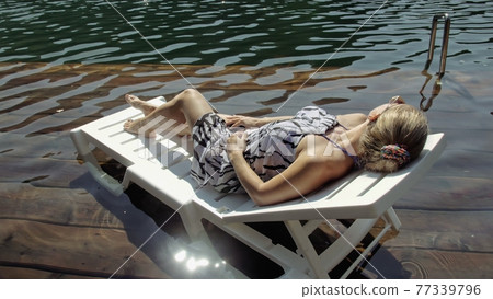 Woman lie on a sunbed in sunglasses and a boho silk shawl. Girl rest on a flood wood underwater pier. The pavement is covered with water in the lake. 77339796