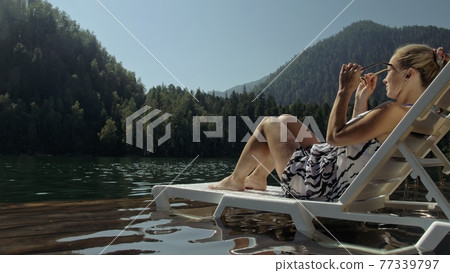 Woman lie on a sunbed in sunglasses and a boho silk shawl. Girl rest on a flood wood underwater pier. The pavement is covered with water in the lake. Woman lie on a sunbed in sunglasses and a boho silk shawl. Girl rest on a flood wood underwater pier. The pavement is covered with water in the lake. 77339797
