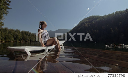 Woman lie on a sunbed in sunglasses and a boho silk shawl. Girl rest on a flood wood underwater pier. The pavement is covered with water in the lake. 77339798