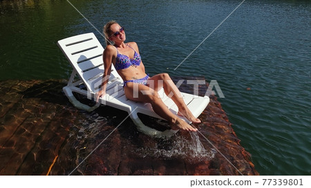 Woman sit on a sunbed in sunglasses and swimming suit. Girl rest on a flood wood underwater pier. The pavement is covered with water in lake. 77339801