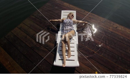 Woman lie on a sunbed in sunglasses and a boho silk shawl. Girl rest on a flood wood underwater pier. The pavement is covered with water in the lake. 77339824