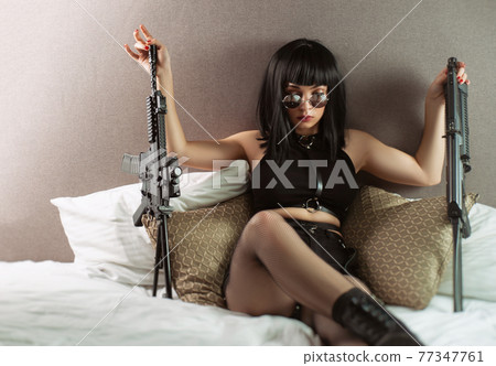 portrait of a female assassin with an automatic rifle in round glasses on the bed 77347761