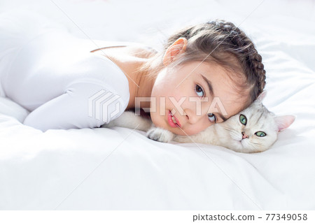 Portrait of a beautiful little girl in white clothes, lies on a clean, white bed with a white British cat. 77349058