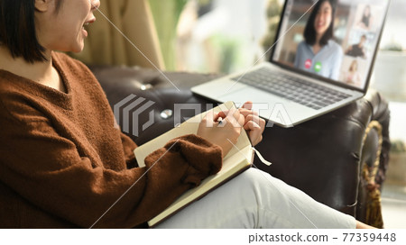Beautiful woman taking notes while sitting in front her laptop at the leather couch in living room. Work from home/Remote working concept. Working from home through video conference concept. Beautiful woman taking notes while sitting in front her laptop at the leather couch in living room. Work from home/Remote working concept. Working from home through video conference concept. 77359448