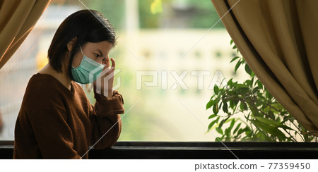 Photo of young woman wearing on medical mask feeling headache while standing at the modern living room windows. Woman keeping a quarantine herself at home. Social distancing concept. 77359450