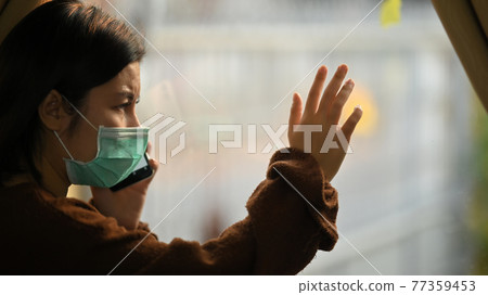 Photo of young woman wearing on medical mask using a smartphone while standing at the modern living room windows. Woman keeping a quarantine herself at home. Social distancing concept. 77359453