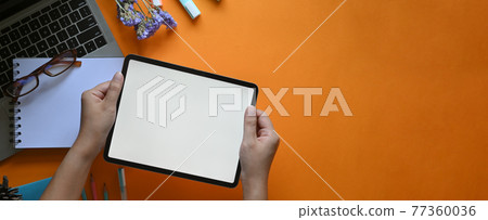 Top view image of creative woman's hands holding a white blank screen computer tablet over computer laptop, glasses, marker pens, pencils, notebook and flowers that putting on colorful working desk. Top view image of creative woman's hands holding a white blank screen computer tablet over computer laptop, glasses, marker pens, pencils, notebook and flowers that putting on colorful working desk. 77360036