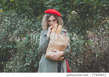 Young woman with wearing red beret holding craft paper bag with french baguette outdoor Young woman with wearing red beret holding craft paper bag with french baguette outdoor 77364631