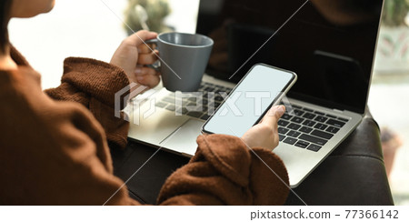 Photo of beautiful woman relaxing on her holidays by sitting at the leather couch and drinking a hot coffee while using a white blank screen computer laptop that putting on her lap. Woman relax time. 77366142