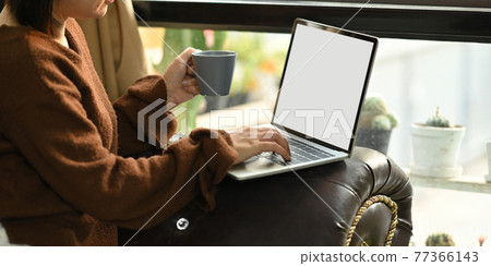 Photo of beautiful woman relaxing on her holidays by sitting at the leather couch and drinking a hot coffee while using a white blank screen computer laptop that putting on her lap. Woman relax time. Photo of beautiful woman relaxing on her holidays by sitting at the leather couch and drinking a hot coffee while using a white blank screen computer laptop that putting on her lap. Woman relax time. 77366143