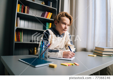 Low-angle view of thinking pupil boy thoughtful writing in notebook with pen sitting at desk near window. Low-angle view of thinking pupil boy thoughtful writing in notebook with pen sitting at desk near window. 77366146
