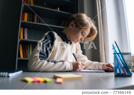 Close-up view of thoughtful smart schoolboy making notes writing essay in notebook sitting at desk. 77366149