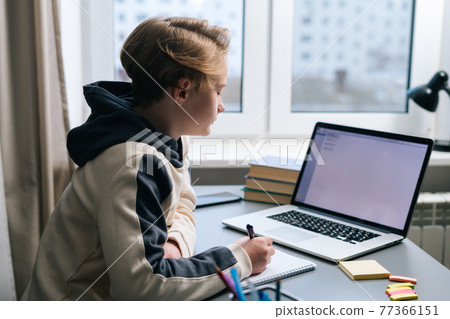 Portrait of pupil boy looking at laptop screen during online lesson and taking notes in copybook. Portrait of pupil boy looking at laptop screen during online lesson and taking notes in copybook. 77366151
