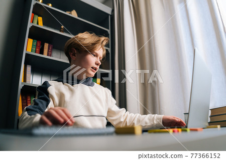 Low-angle view of pupil boy using typing laptop computer doing online lesson via Internet sitting at desk, selective focus. Low-angle view of pupil boy using typing laptop computer doing online lesson via Internet sitting at desk, selective focus. 77366152