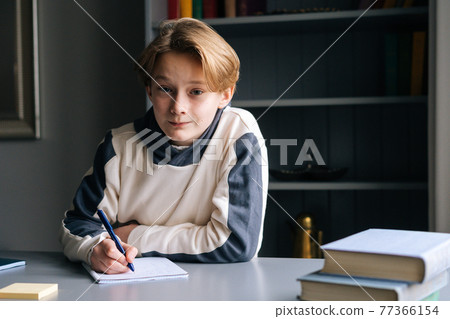 Portrait of smiling pupil boy doing homework writing to notebook with pen sitting at desk on background of book shelves and looking at camera. Portrait of smiling pupil boy doing homework writing to notebook with pen sitting at desk on background of book shelves and looking at camera. 77366154