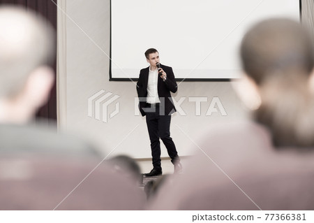 speaker conducts the business of the conference standing in front of a large white screen on the stage in the conference room 77366381