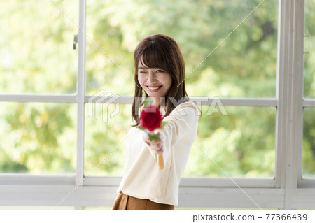 一個年輕女子在她面前的窗邊拿著一朵玫瑰 一個年輕女子在她面前的窗邊拿著一朵玫瑰 77366439