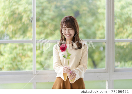 一個年輕女子在窗邊拿著一朵玫瑰 一個年輕女子在窗邊拿著一朵玫瑰 77366451