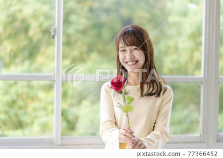 一個年輕女子在窗邊拿著一朵玫瑰 一個年輕女子在窗邊拿著一朵玫瑰 77366452