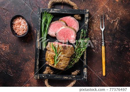 Roast beef round fillet meat in a wooden tray with herbs. Dark background. Top view Roast beef round fillet meat in a wooden tray with herbs. Dark background. Top view 77376497