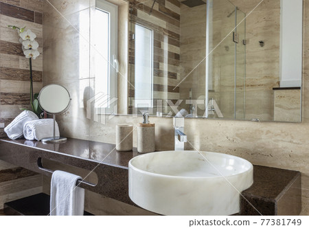 Elegant and contemporary looking sink with a mirror in a brown colored and bright bathroom. It is decorated with flowers and some towels. Elegant and contemporary looking sink with a mirror in a brown colored and bright bathroom. It is decorated with flowers and some towels. 77381749