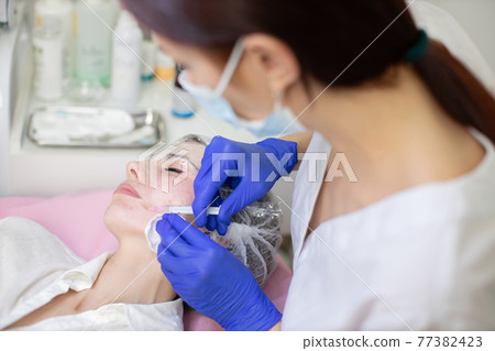 Beautiful woman at the dermatology clinic. Cosmetologist doing injections on the face and mouth area of female client. Skin biorevitalizing and anti-aging therapy. Beautiful woman at the dermatology clinic. Cosmetologist doing injections on the face and mouth area of female client. Skin biorevitalizing and anti-aging therapy. 77382423