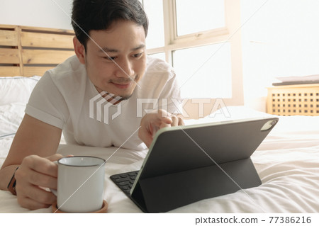 Happy man works on his tablet with a cup of coffee on the bed. Happy man works on his tablet with a cup of coffee on the bed. 77386216