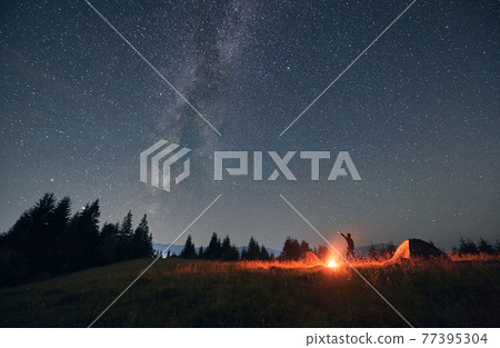 Male traveler standing near campfire and pointing at stars. Male traveler standing near campfire and pointing at stars. 77395304