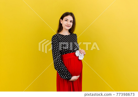 Happy pregnant woman holding baby girl booties against her belly at Colored background. Happy pregnancy. Copy space 77398673