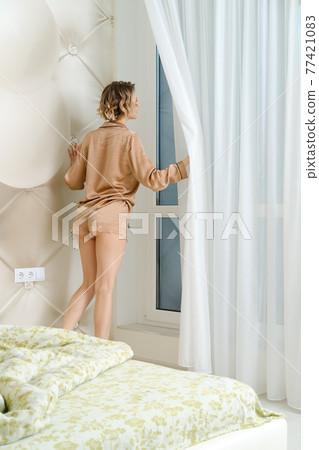 Woman in pajamas gets out of bed, comes to the window and moves the curtain aside 77421083