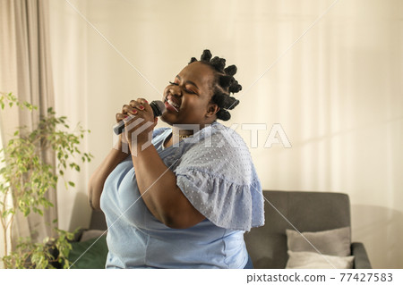 Woman holding microphone and singing a song at home 77427583