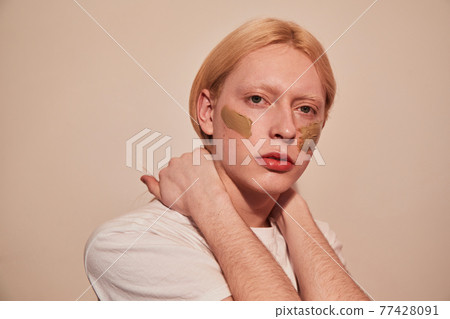 Feminine man posing in studio with concealer at his chick isolated on a beige background Feminine man posing in studio with concealer at his chick isolated on a beige background 77428091