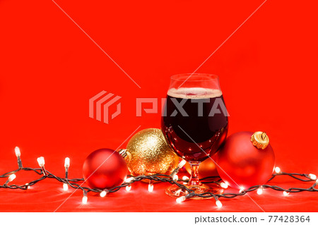 Dark ale beer in a tulip glass with christmas baubles and lights on red background Dark ale beer in a tulip glass with christmas baubles and lights on red background 77428364
