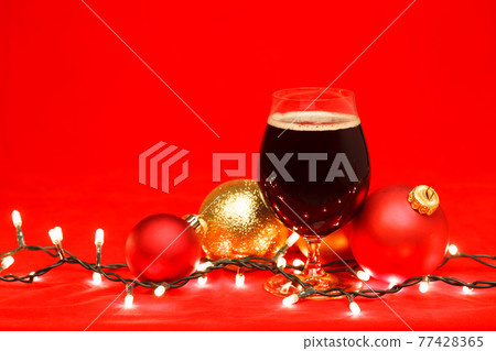 Dark ale beer in a tulip glass with christmas baubles and lights on red background 77428365
