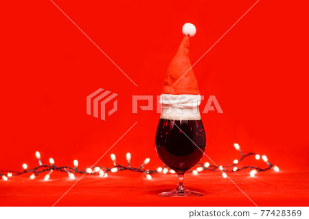 Dark ale beer in a tulip glass with red santa hat and christmas lights on red background Dark ale beer in a tulip glass with red santa hat and christmas lights on red background 77428369