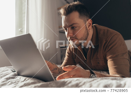 Man laying at the stomach at the bed in front of the laptop and working 77428905