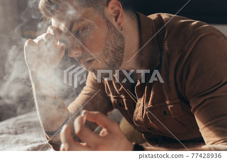 Man blows smoke while smoking marijuana hand rolled cigarette and sitting Man blows smoke while smoking marijuana hand rolled cigarette and sitting 77428936