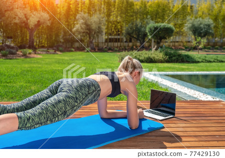 Woman is practicing fitness gymnastics, pilates, on outdoor near house and pool, online lessons with a trainer. With a background to garden. Woman is practicing fitness gymnastics, pilates, on outdoor near house and pool, online lessons with a trainer. With a background to garden. 77429130
