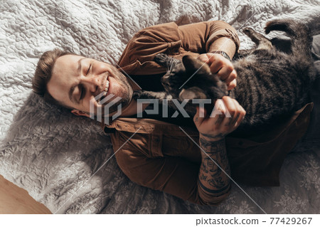 Man laughing and stroking his striped cat while spending time with him at bedroom 77429267
