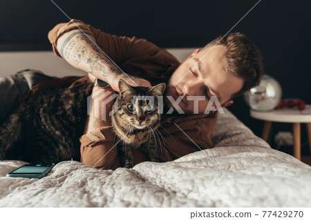 Man stroking his striped cat while spending time with him at the bedroom 77429270