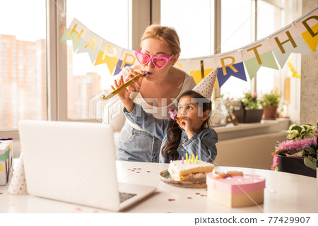 Mother and her little funny daughter blowing a pipe and looking at the laptop screen 77429907
