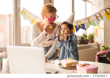Mother and her little funny daughter blowing a pipe and looking at laptop screen 77429917