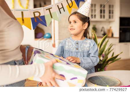 Girl wearing party hat looking at her mother while accepting her birthday gift 77429927