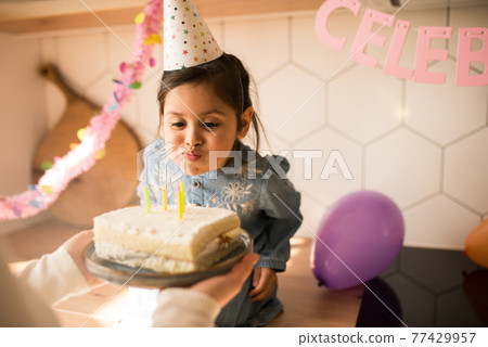 Girl wearing party cap blowing on the candles at her birthday cake Girl wearing party cap blowing on the candles at her birthday cake 77429957
