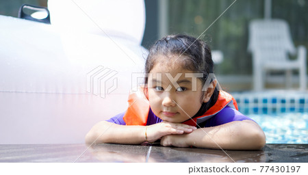 Asian child playing in the pool. Wearing orange life jacket, rests her chin on both hands by the edge of the pool. Asian child playing in the pool. Wearing orange life jacket, rests her chin on both hands by the edge of the pool. 77430197