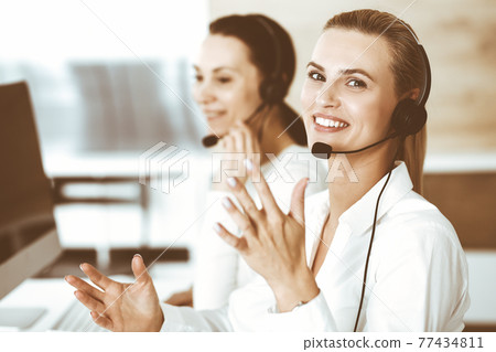 Blond female customer service representative and her colleague are consulting clients online using headset. Call center and business people concept Blond female customer service representative and her colleague are consulting clients online using headset. Call center and business people concept 77434811
