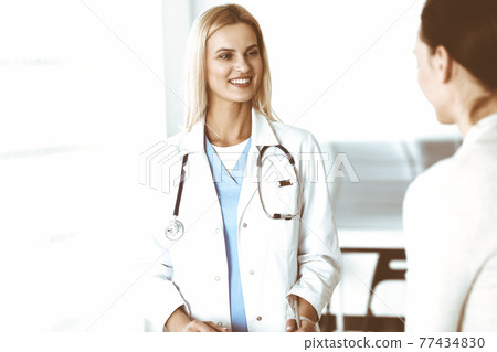 Woman-doctor at work in hospital is happy to communicate with female patient. Blonde physician checks medical history record and exam results. Medicine concept 77434830