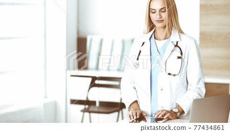 Woman-doctor at work in clinic excited and happy of her profession. Blond female physician is smiling while using tablet computer. Medicine concept Woman-doctor at work in clinic excited and happy of her profession. Blond female physician is smiling while using tablet computer. Medicine concept 77434861